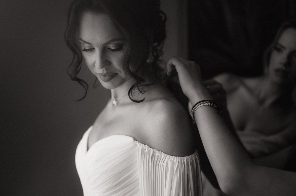 Black and white photo of a bride in a strapless wedding gown adjusting her earring with a mirror reflection behind her.