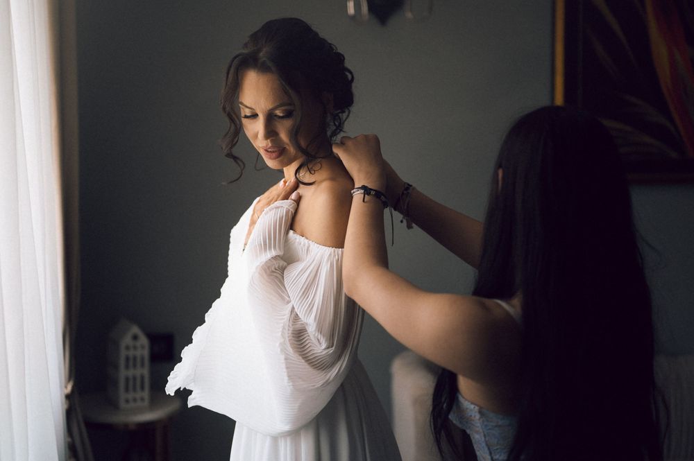 Bridesmaid helping bride put on white wedding dress in a softly lit room.