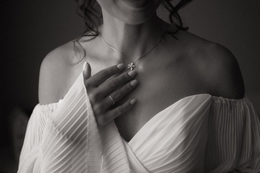 Black and white photo of a bride in white off-shoulder gown, hands clasped at chest, wearing a delicate necklace.