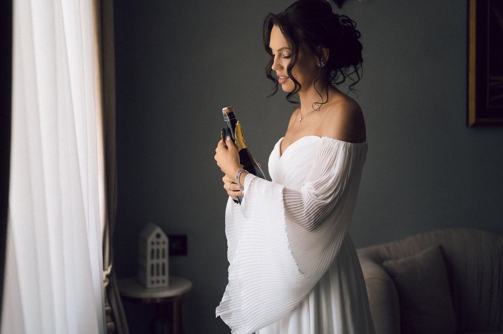 Bride in white off-shoulder gown holding champagne bottle by window on wedding day.