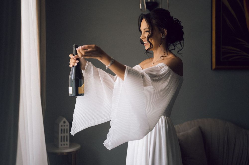 Bride in white off-shoulder gown smiling while opening champagne bottle indoors.