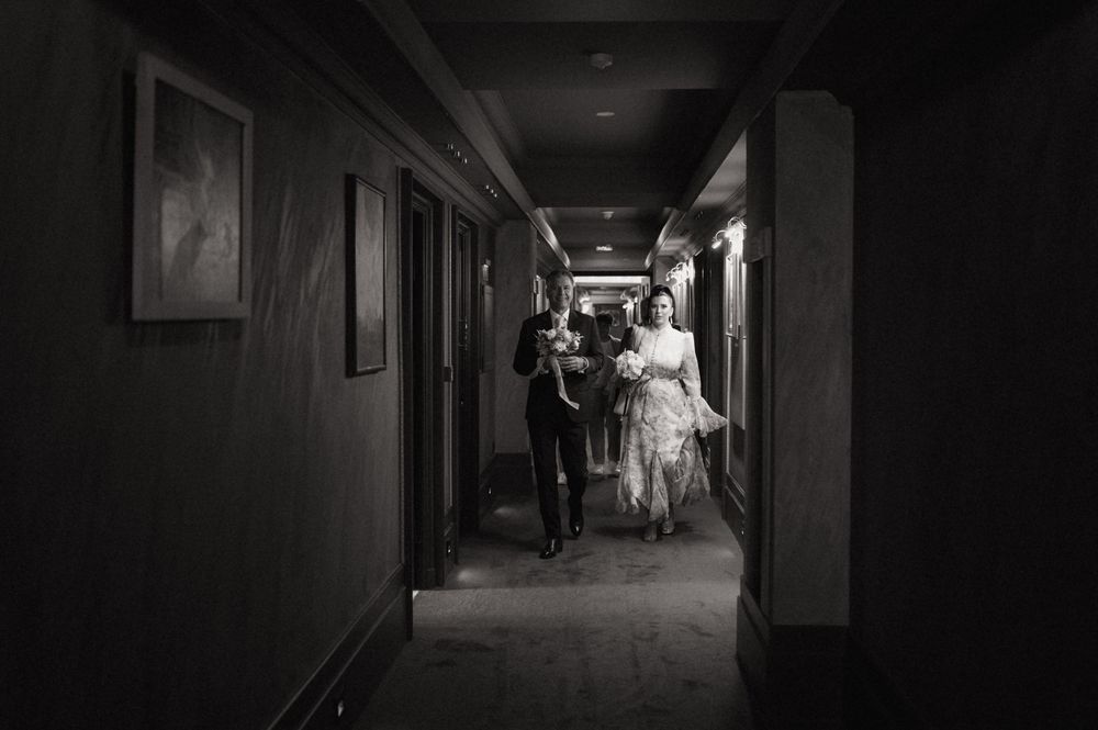 Black and white photo of a bride and groom walking down a dimly lit hotel hallway on their wedding day.