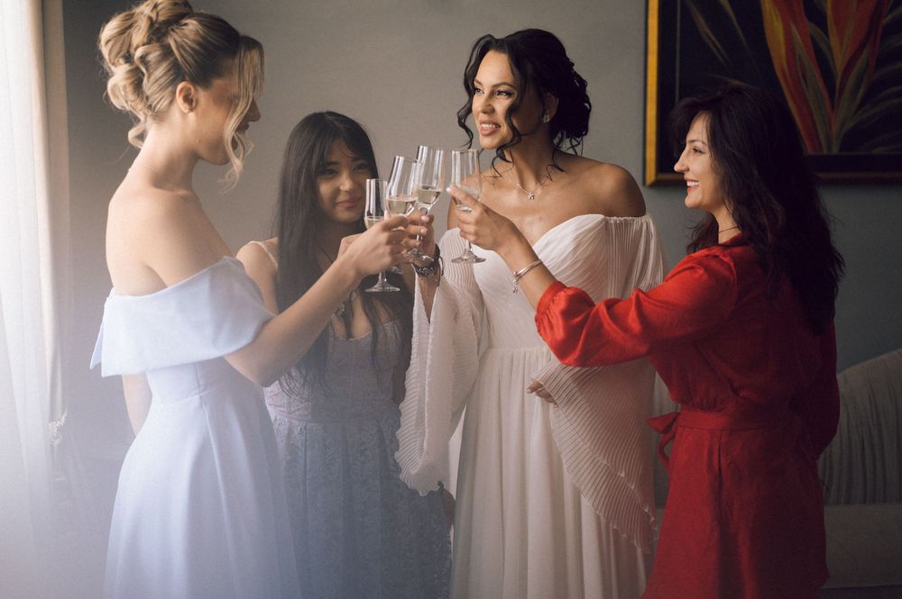 Bride and bridesmaids in white dresses toasting with champagne glasses, celebrating wedding day indoors.