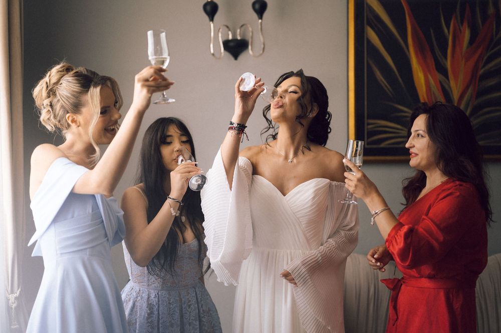 Bride in white gown toasts with three bridesmaids, one in blue dress and one in red, celebrating before the wedding.