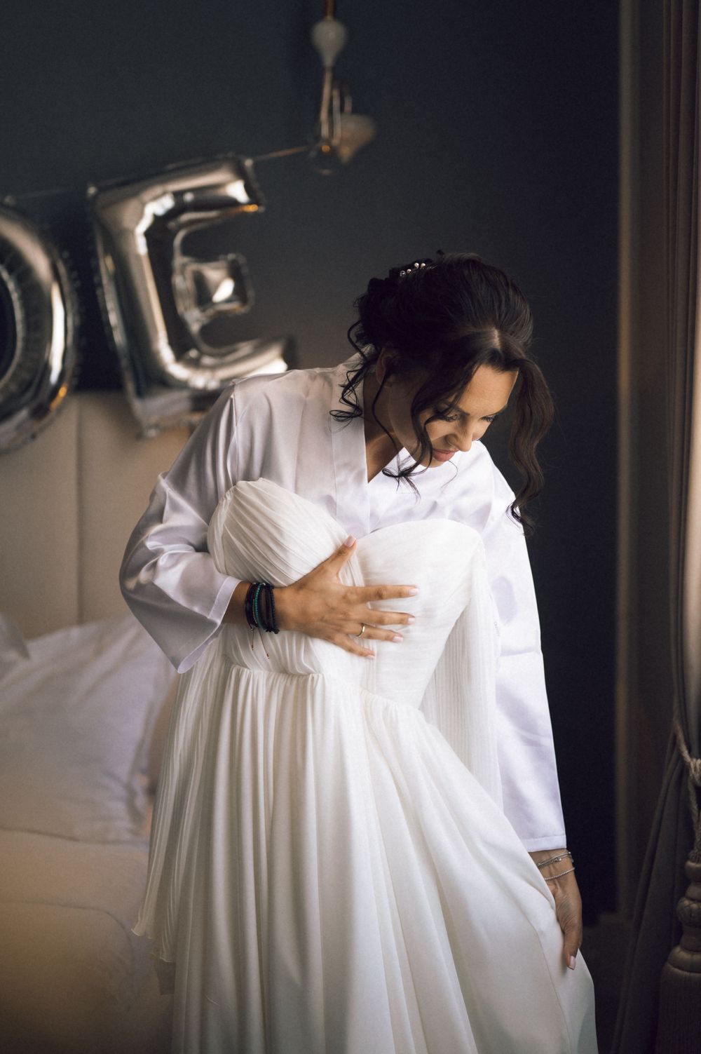 Bride in white robe holding her wedding dress near silver LOVE balloons in a softly lit bedroom.