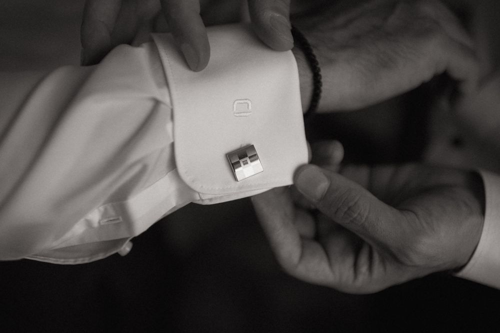 Black and white photo of hands fastening a square cufflink on a white dress shirt sleeve.
