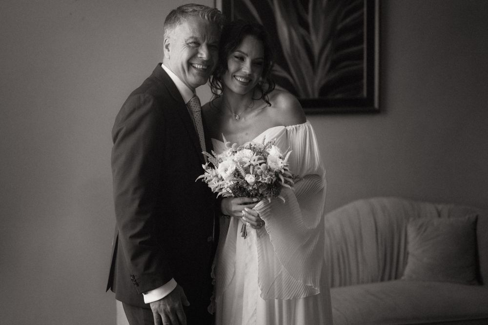 Black and white wedding photo of smiling bride holding bouquet and groom in suit posing together indoors.