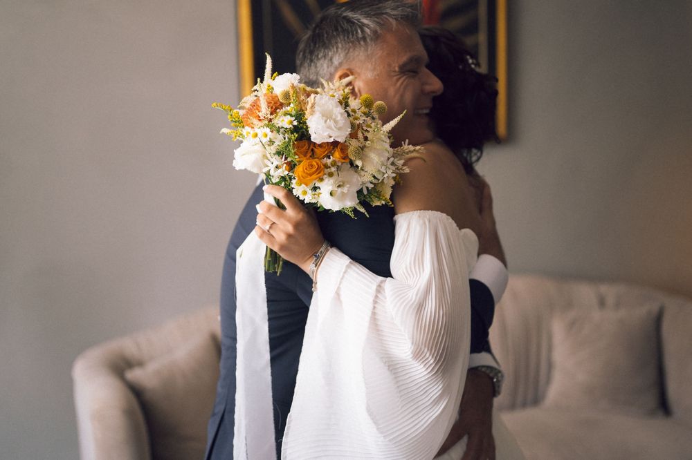 Couple embracing at wedding, bride holding colorful bouquet of white and orange flowers in elegant room.