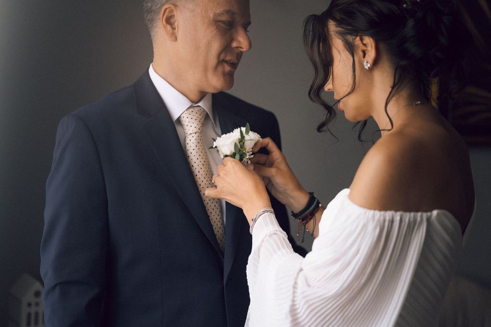 A bride in a white off-shoulder dress pins a white boutonniere on a groom's navy suit lapel.