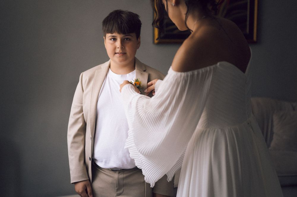 A bride in a white dress pins a boutonniere on a young boy in a tan suit during wedding preparations.