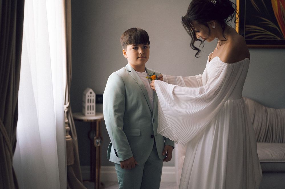 Bride in white dress pinning boutonniere on young boy in light blue suit indoors.