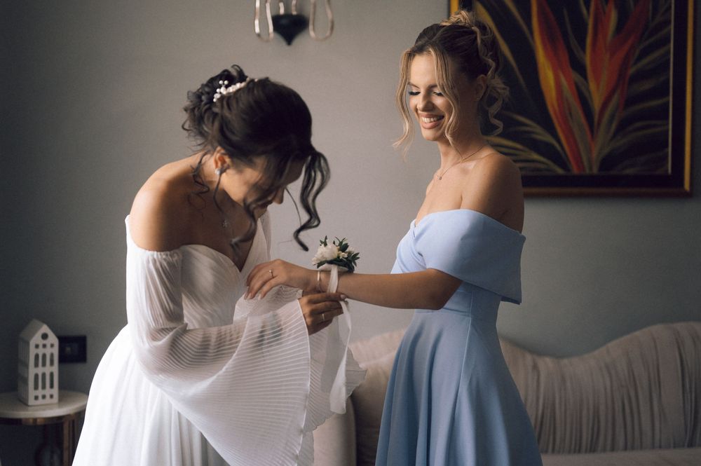 Bride in white dress getting corsage pinned by smiling bridesmaid in light blue off-shoulder gown indoors.