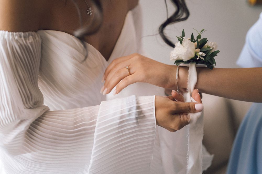 Bride helping bridesmaid put on a white floral wrist corsage with ribbon during wedding preparations.