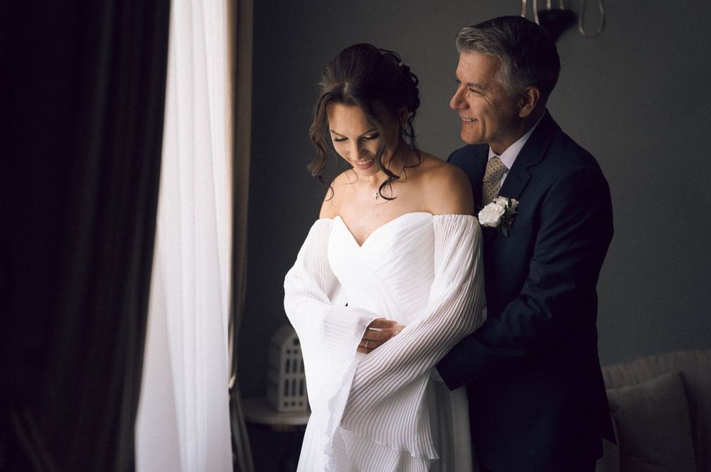Groom tenderly embraces bride in white off-shoulder wedding dress near a window in elegant indoor setting.