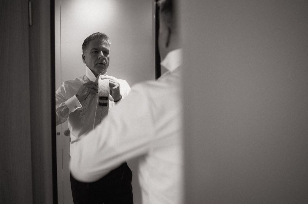Black and white photo of a man adjusting his tie in a mirror while getting dressed in a white shirt.