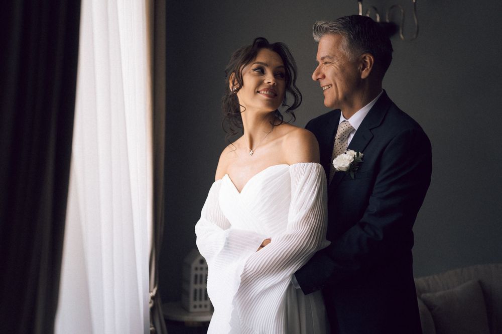 Elegant bride and groom embracing by a window on their wedding day, bride wearing off-shoulder white gown.