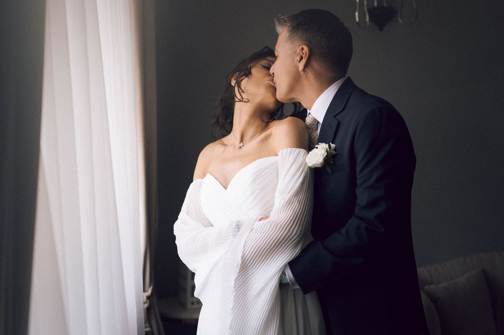 A bride and groom share a romantic kiss near a window, dressed in elegant wedding attire.