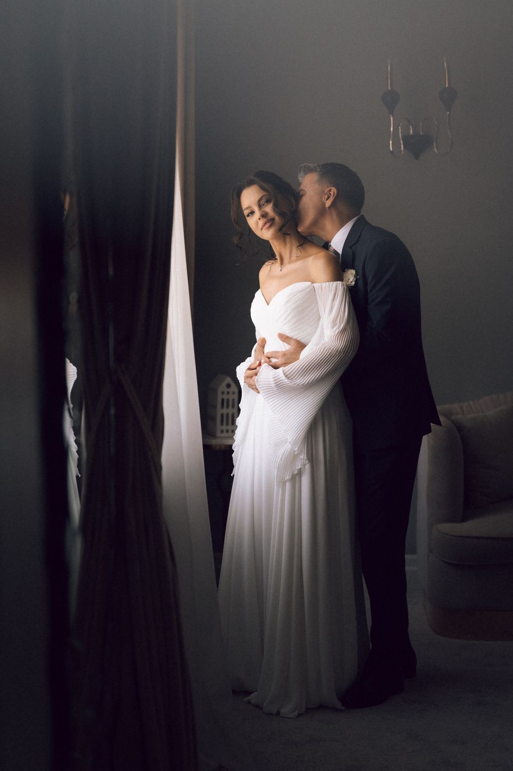 Elegant bride in white off-shoulder gown embraced by groom in dark suit sharing a tender moment indoors.