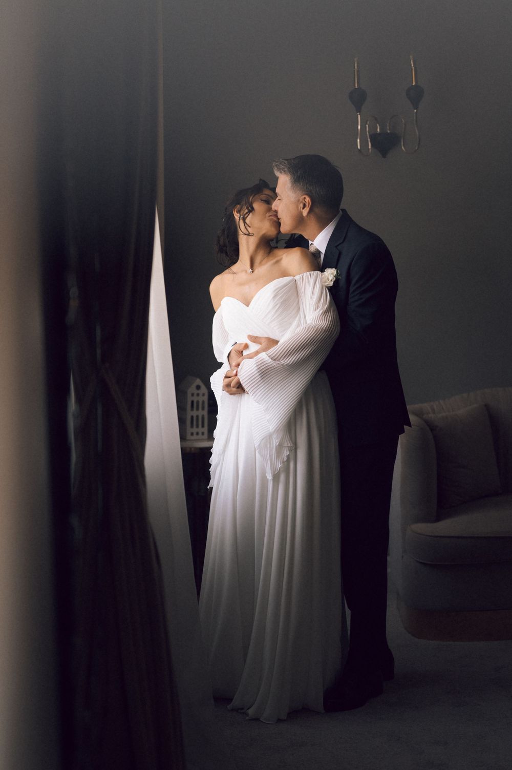 Elegant bride in white off-shoulder gown shares a romantic kiss with groom in black tuxedo in a dimly lit room.