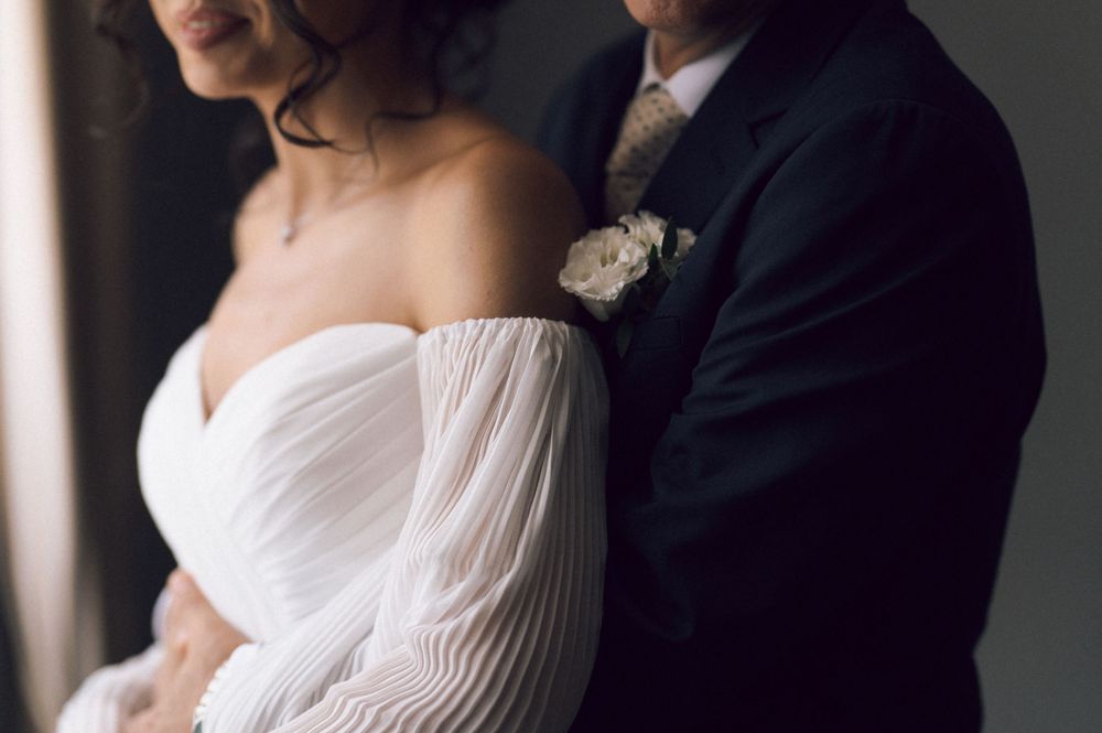 Bride in off-shoulder white gown with groom in dark suit and boutonniere at wedding ceremony.