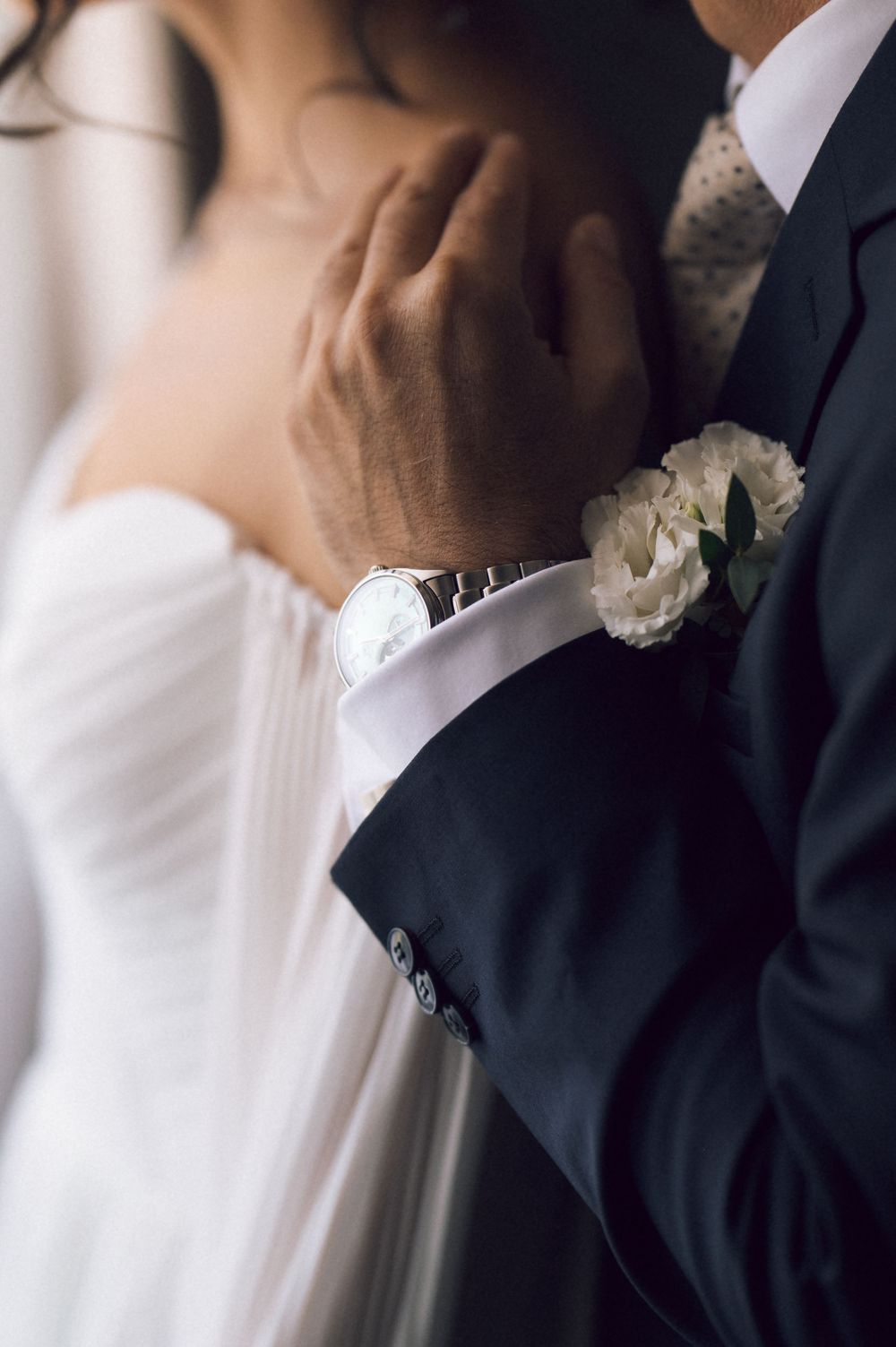 Close-up of groom's hand with silver watch and white boutonniere gently touching bride's neck at wedding.