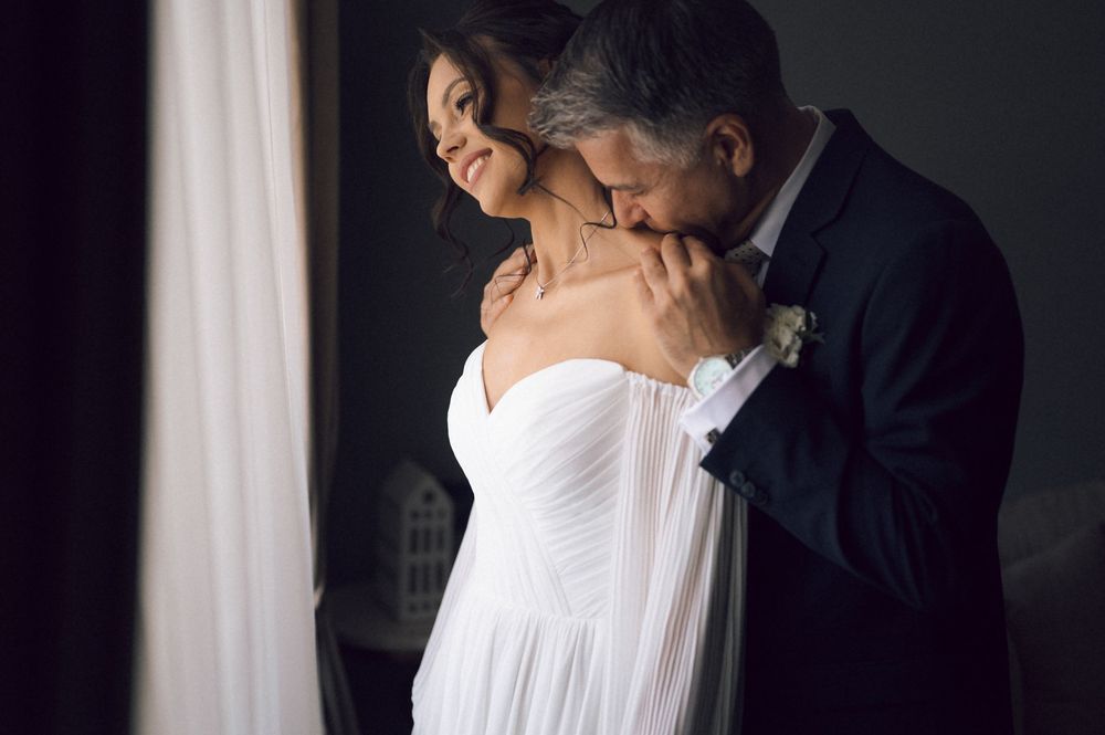 Bride in white strapless gown embraced by groom in black tuxedo near window in romantic wedding portrait.