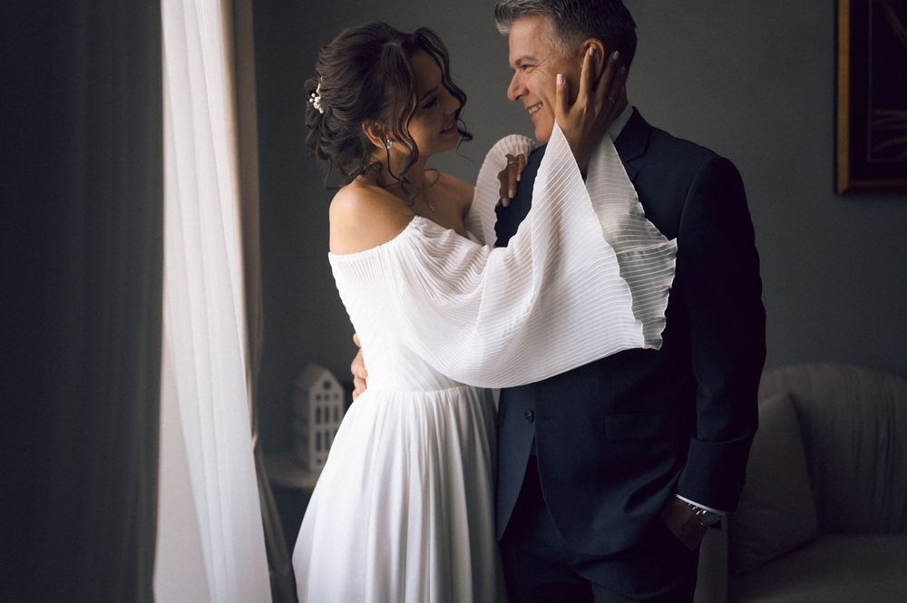 Bride in white off-shoulder gown tenderly touches groom's face as they share an intimate moment indoors.