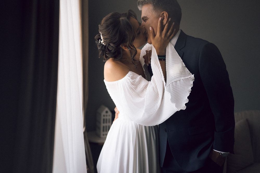 A bride and groom share a romantic kiss near a window, dressed in elegant wedding attire.