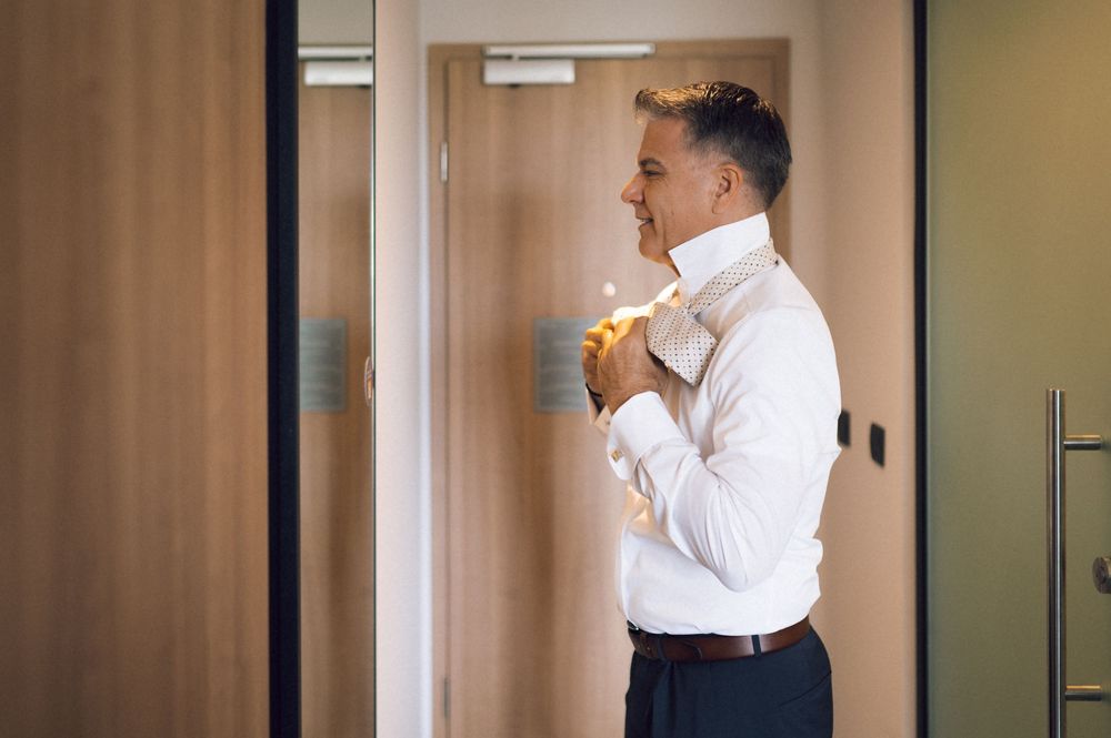 Man in white dress shirt adjusting his collar while getting ready, smiling in a modern room.