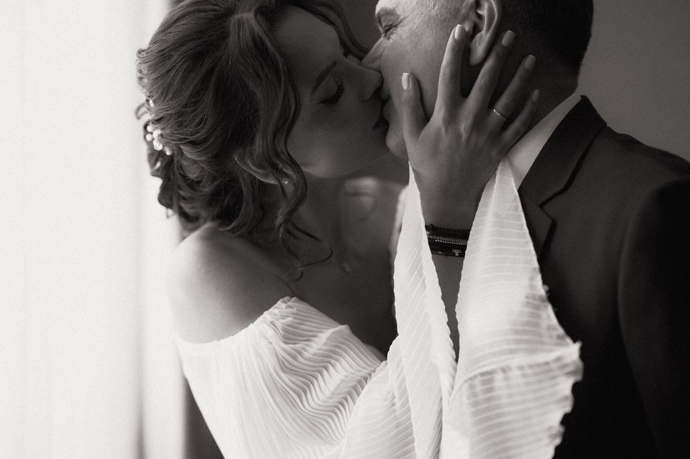 Black and white photo of a couple sharing a romantic kiss, bride in off-shoulder gown and groom in dark suit.