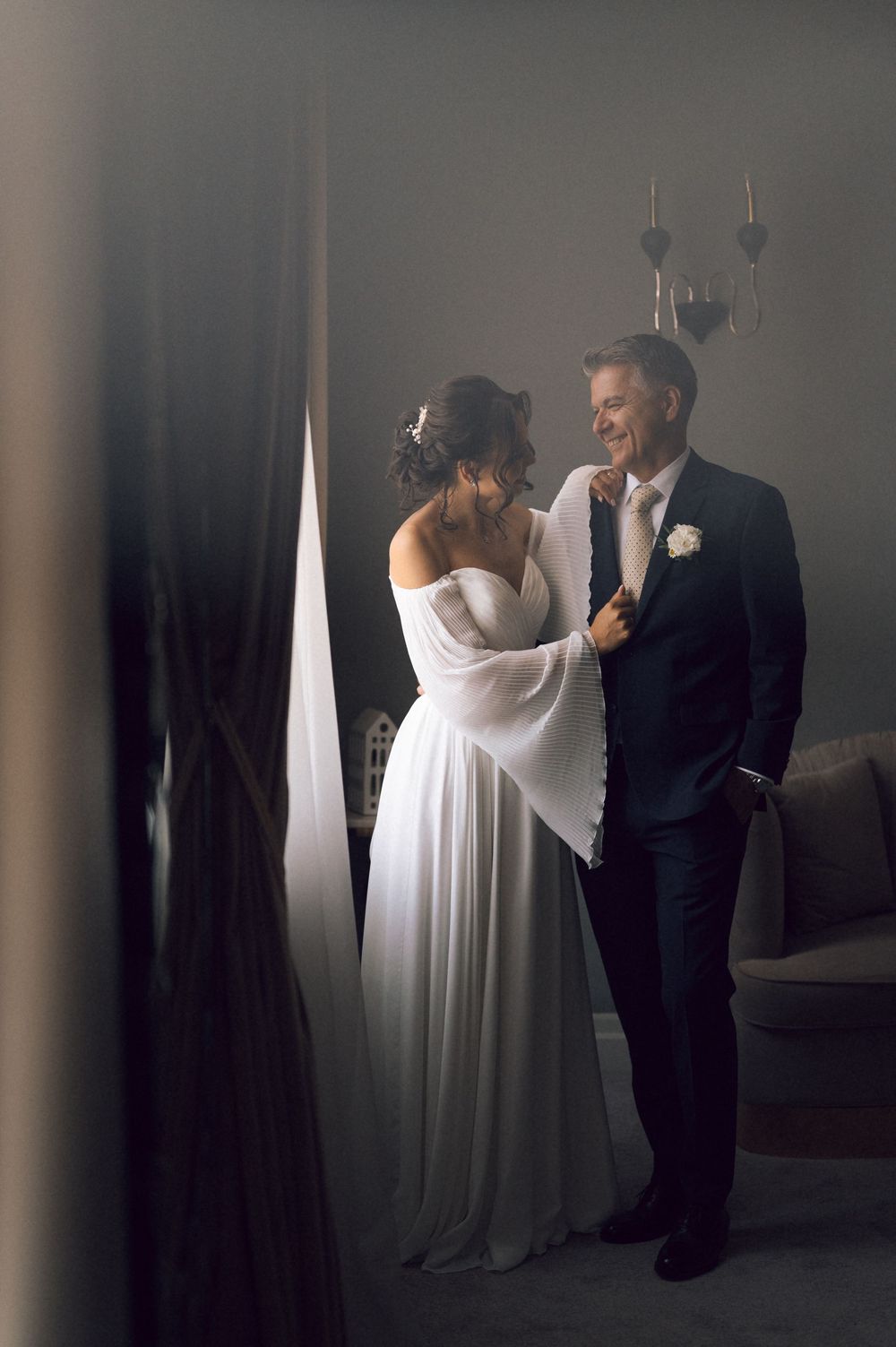Bride in white gown adjusts groom's tie as they share a tender moment by a window on their wedding day.