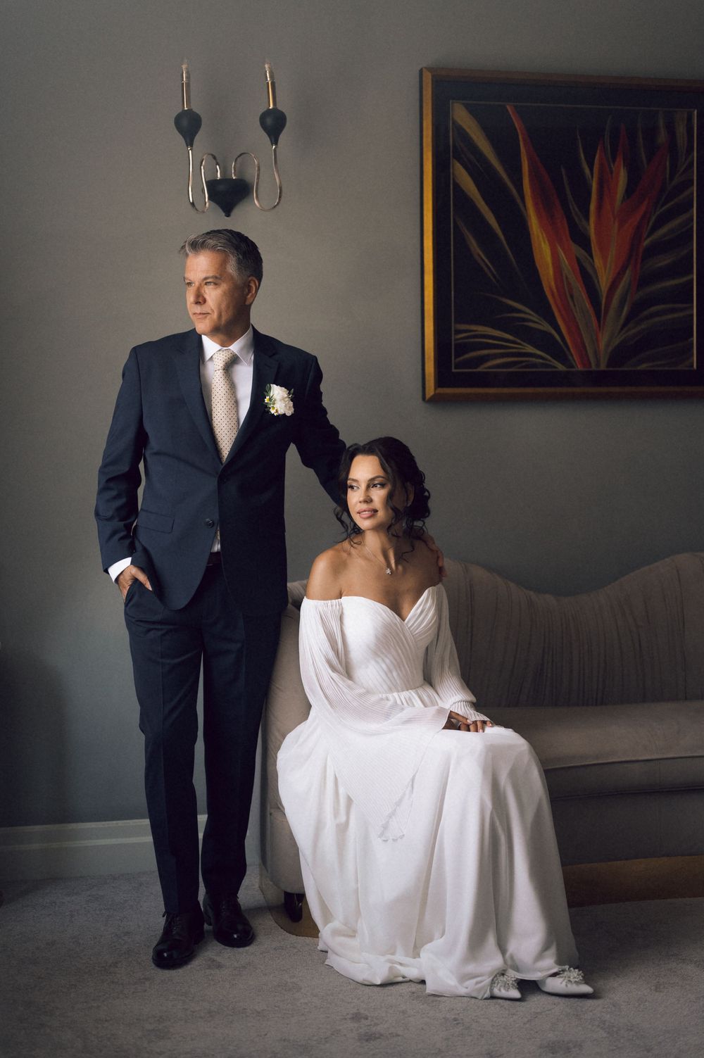 Elegant wedding couple posing indoors, groom in navy suit standing beside seated bride in white off-shoulder gown.