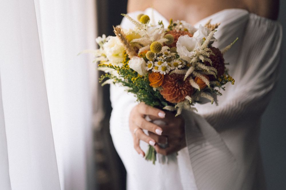 Bride in white off-shoulder gown holding rustic autumn bouquet with orange mums, white blooms, and yellow craspedia.