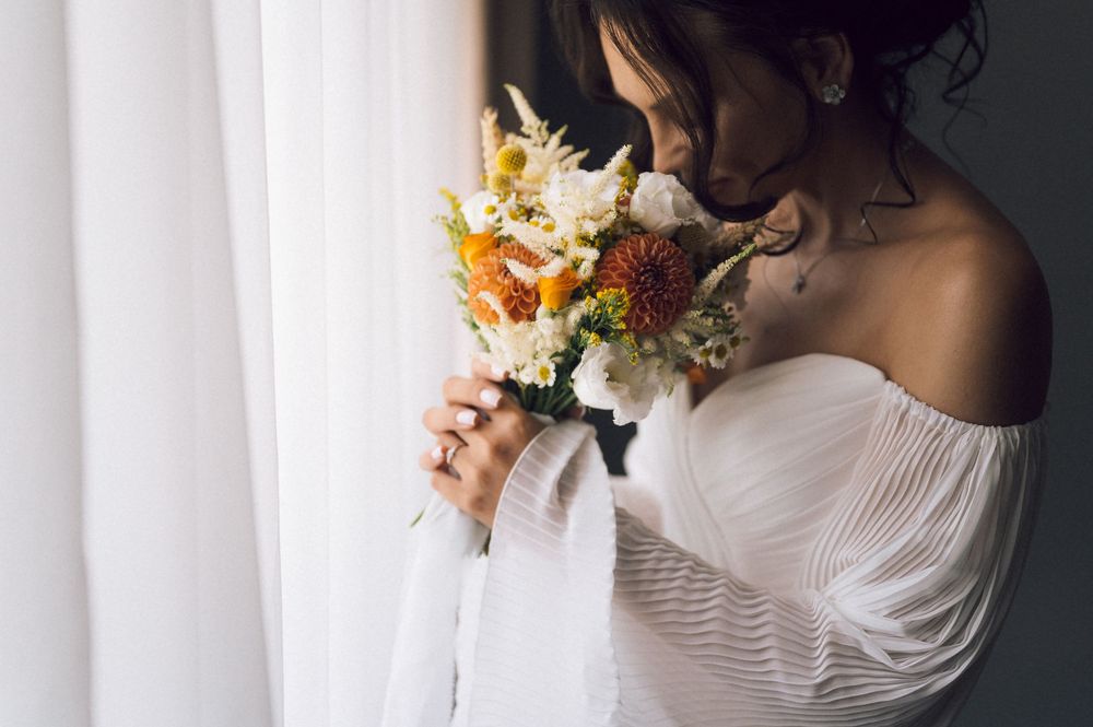 Bride in white off-shoulder dress smelling colorful wedding bouquet with orange and white flowers by a window.
