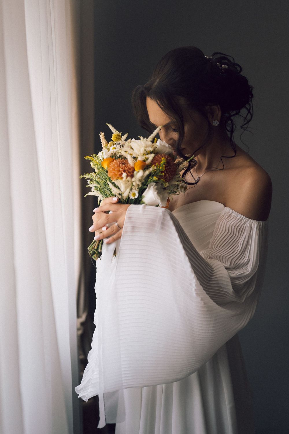 Bride in off-shoulder white gown smelling colorful wedding bouquet with orange and white flowers near curtained window.