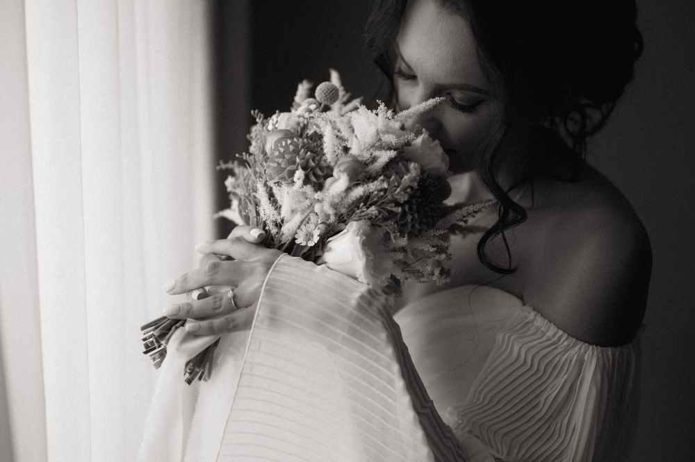 Black and white photo of a bride smelling her wedding bouquet near a window in her bridal gown.