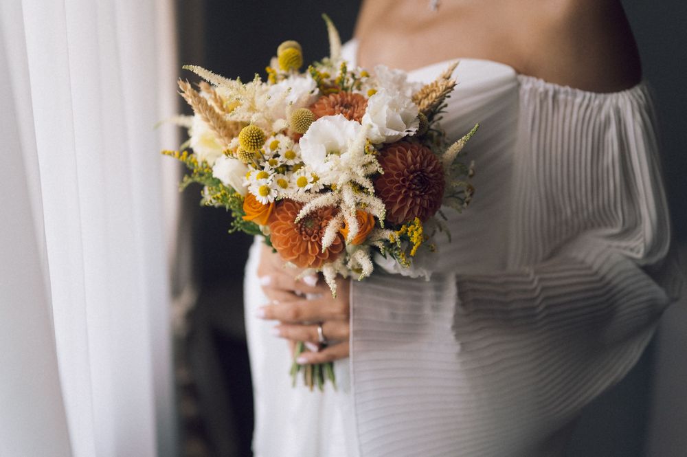 Bride in off-shoulder white dress holding a rustic bouquet with orange, white, and yellow dried flowers.