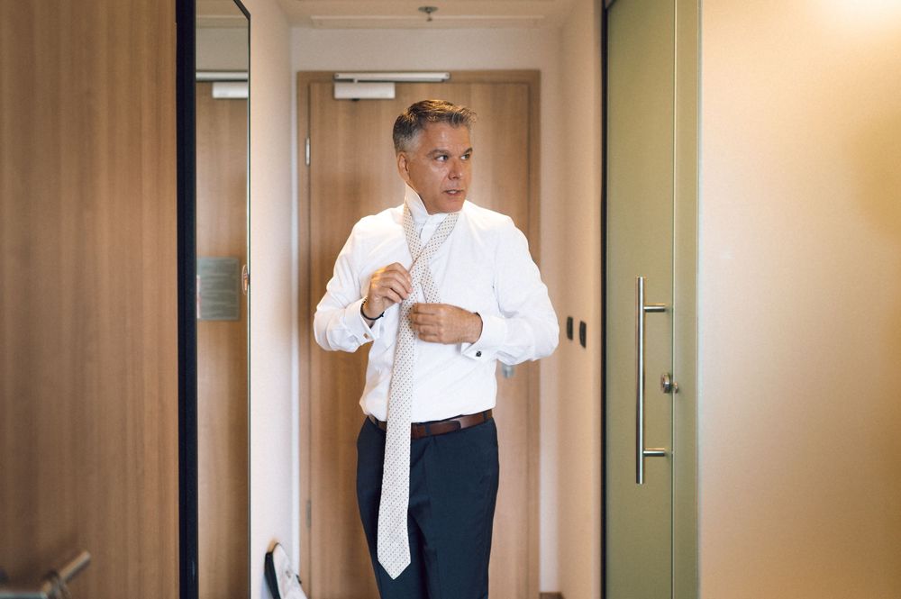 Man in white dress shirt adjusting tie while getting ready in a hotel room doorway.