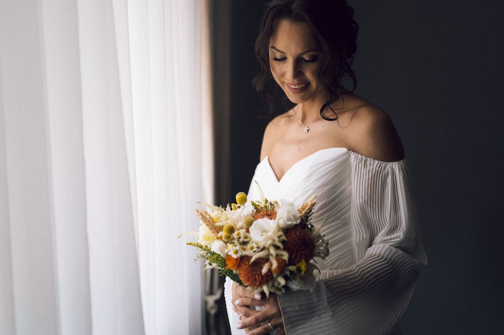 Bride in off-shoulder white gown holding a rustic bouquet of white and orange flowers by a window.
