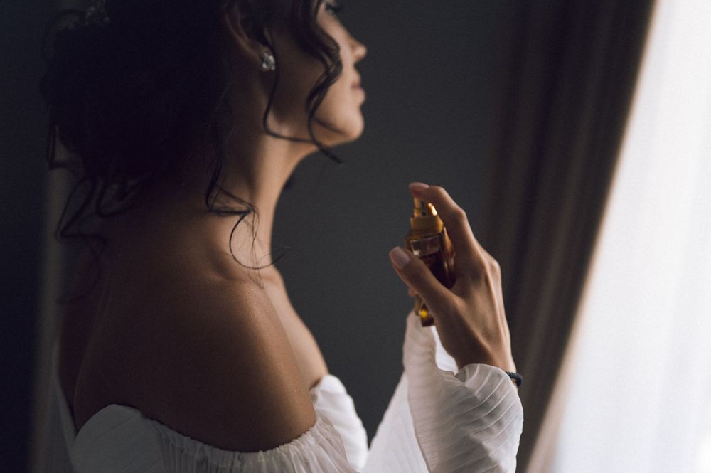Woman in off-shoulder dress applying perfume from a glass bottle, captured in moody, artistic side profile.