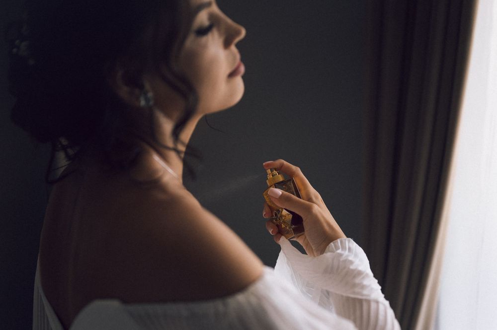 Elegant woman in white dress applying perfume to her neck near a window in soft natural light.