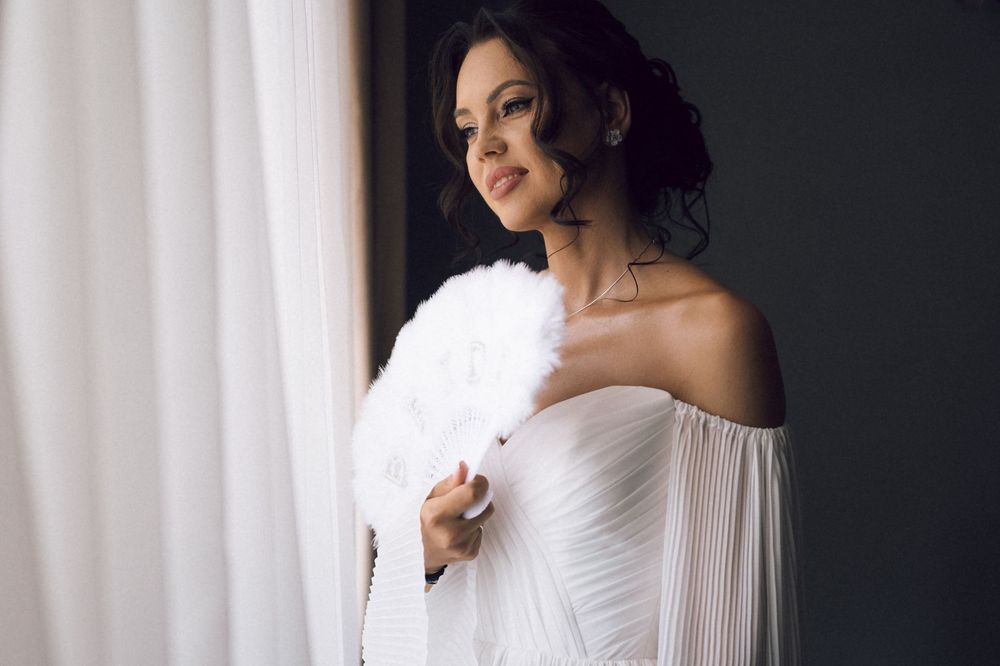 Elegant woman in white off-shoulder dress holding a feather fan near a curtained window.