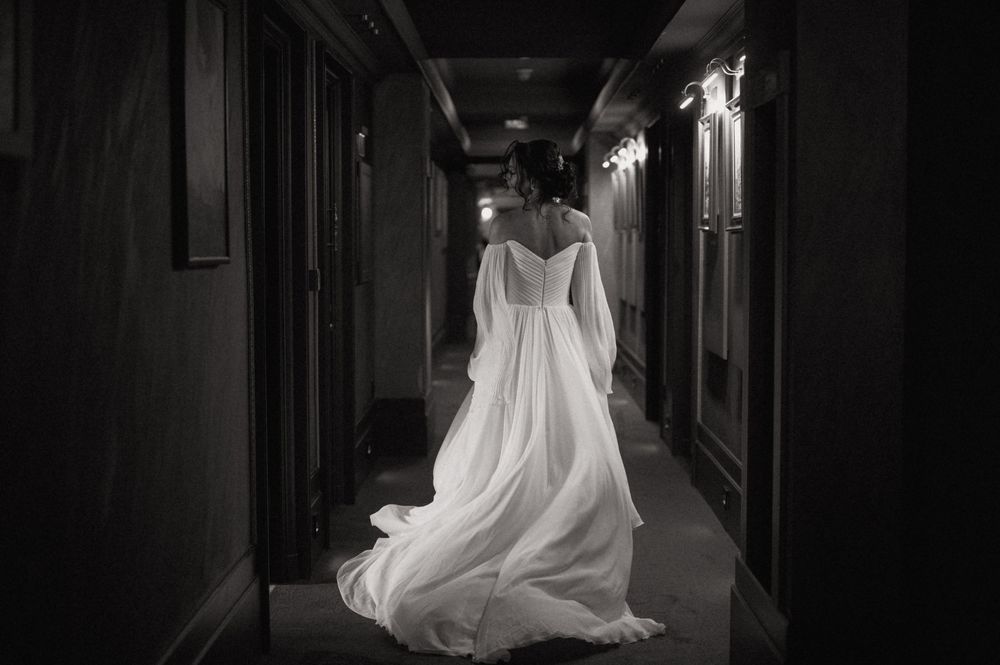 Black and white photo of a bride in a flowing white gown walking down an elegant hallway, her long train trailing behind.