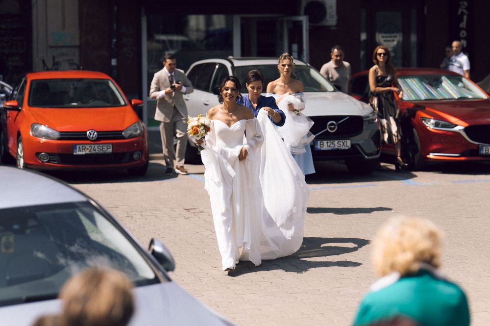 Two brides in white gowns walk through a street lined with cars on a sunny wedding day.