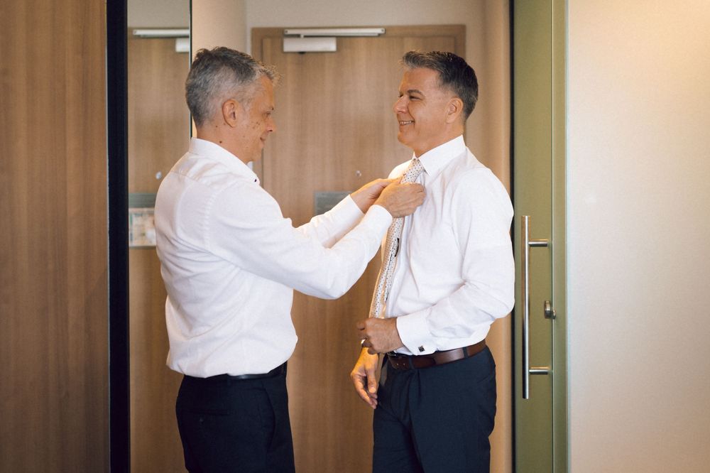 Two men in white dress shirts, one adjusting the other's collar, smiling in a modern room.