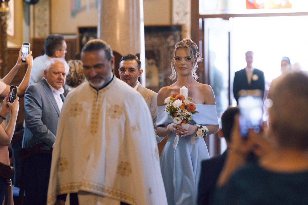 Bride in blue gown walks down church aisle with priest in white vestments during Orthodox wedding ceremony.