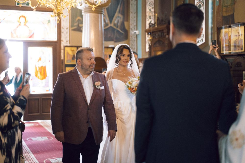 Bride in white gown walks down church aisle with escort during traditional wedding ceremony.