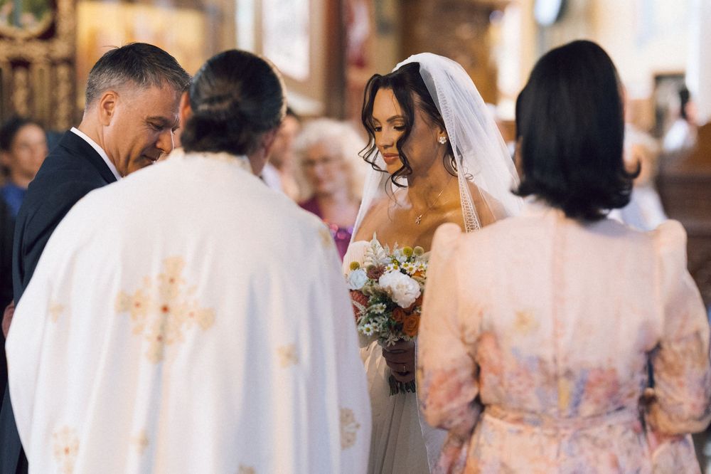 Bride in white gown and veil holds bouquet during Orthodox church wedding ceremony with priest and guests.