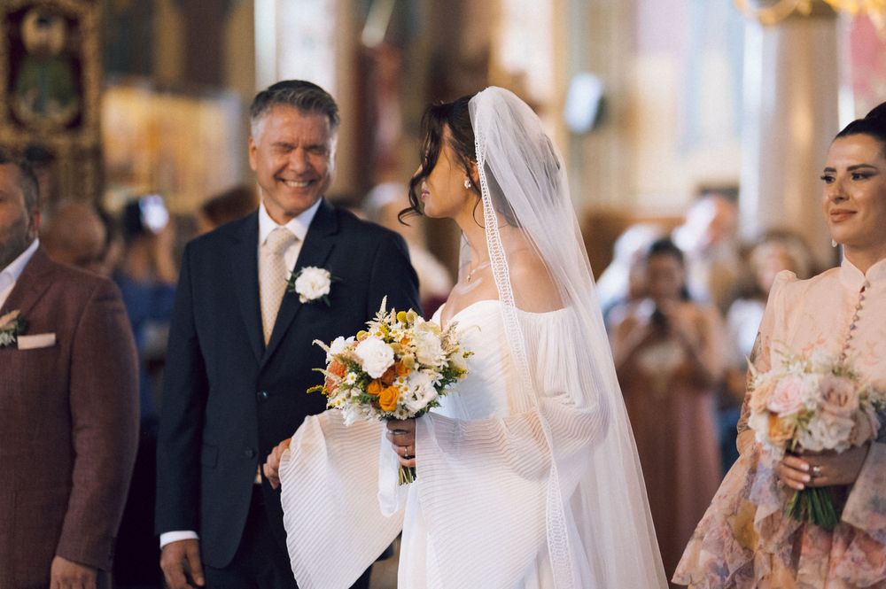 Bride in white gown and veil holds floral bouquet while walking down church aisle with smiling man in suit.
