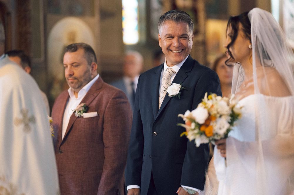 Smiling groom in navy suit stands at church altar with bride in white veil holding orange and white bouquet.
